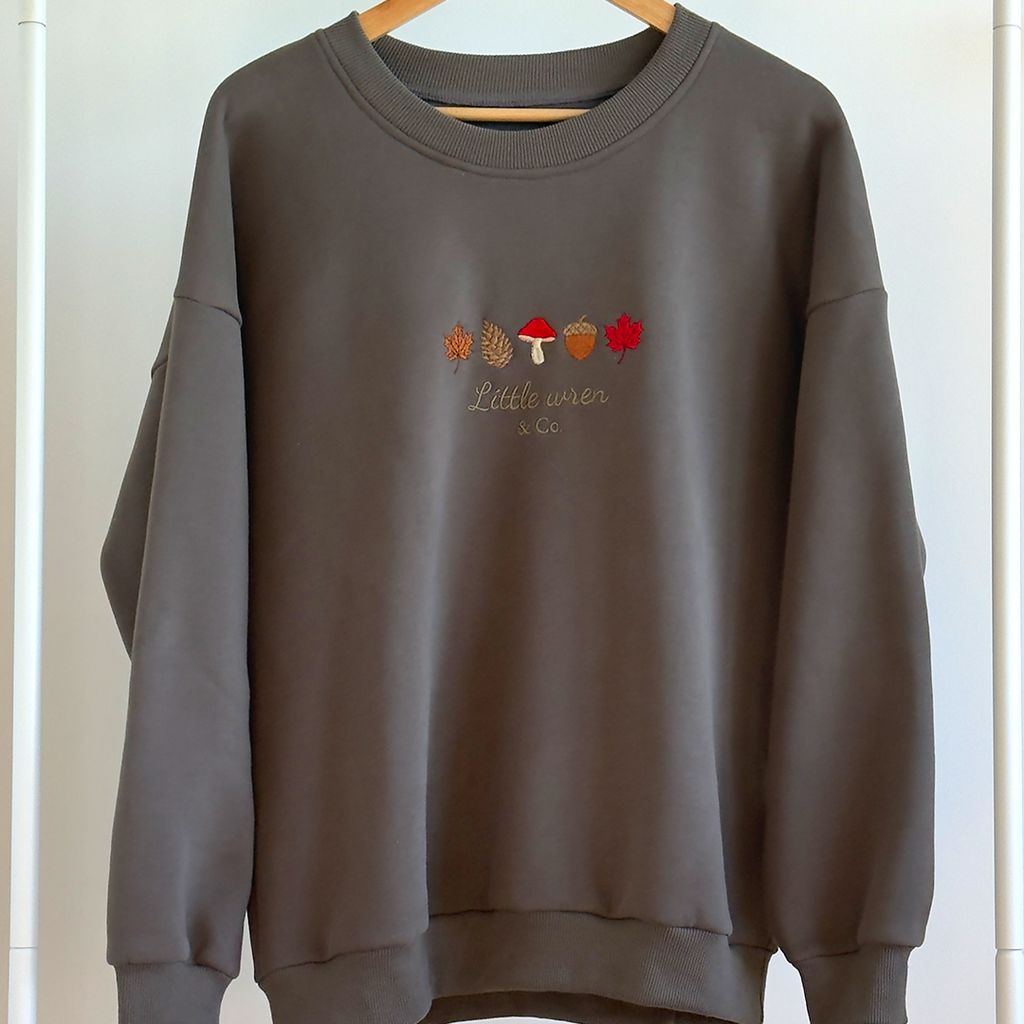 Dark Green sweatshirt with autumn-themed design on a wooden hanger against a white wall.