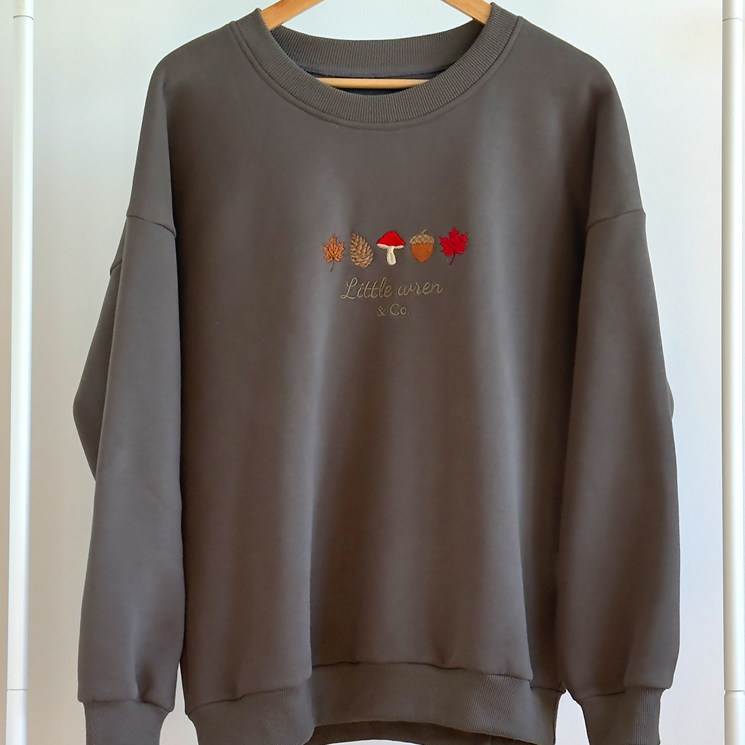 Dark Green sweatshirt with autumn-themed design on a wooden hanger against a white wall.