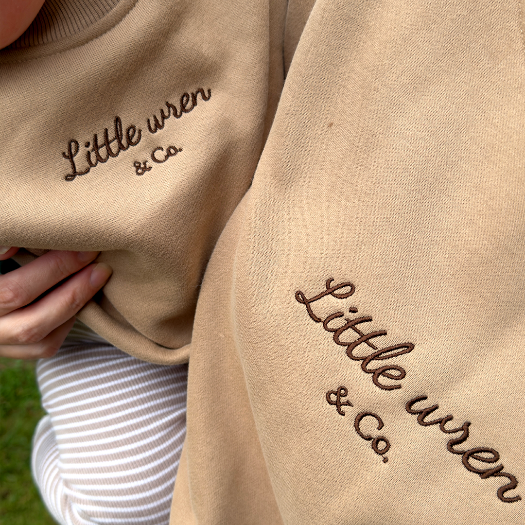 Beige sweatshirt with 'Little wren & co.' embroidery on a grassy background