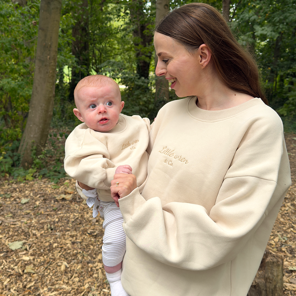 Woman holding a baby in a forest setting wearing matching cream sweatshirts.