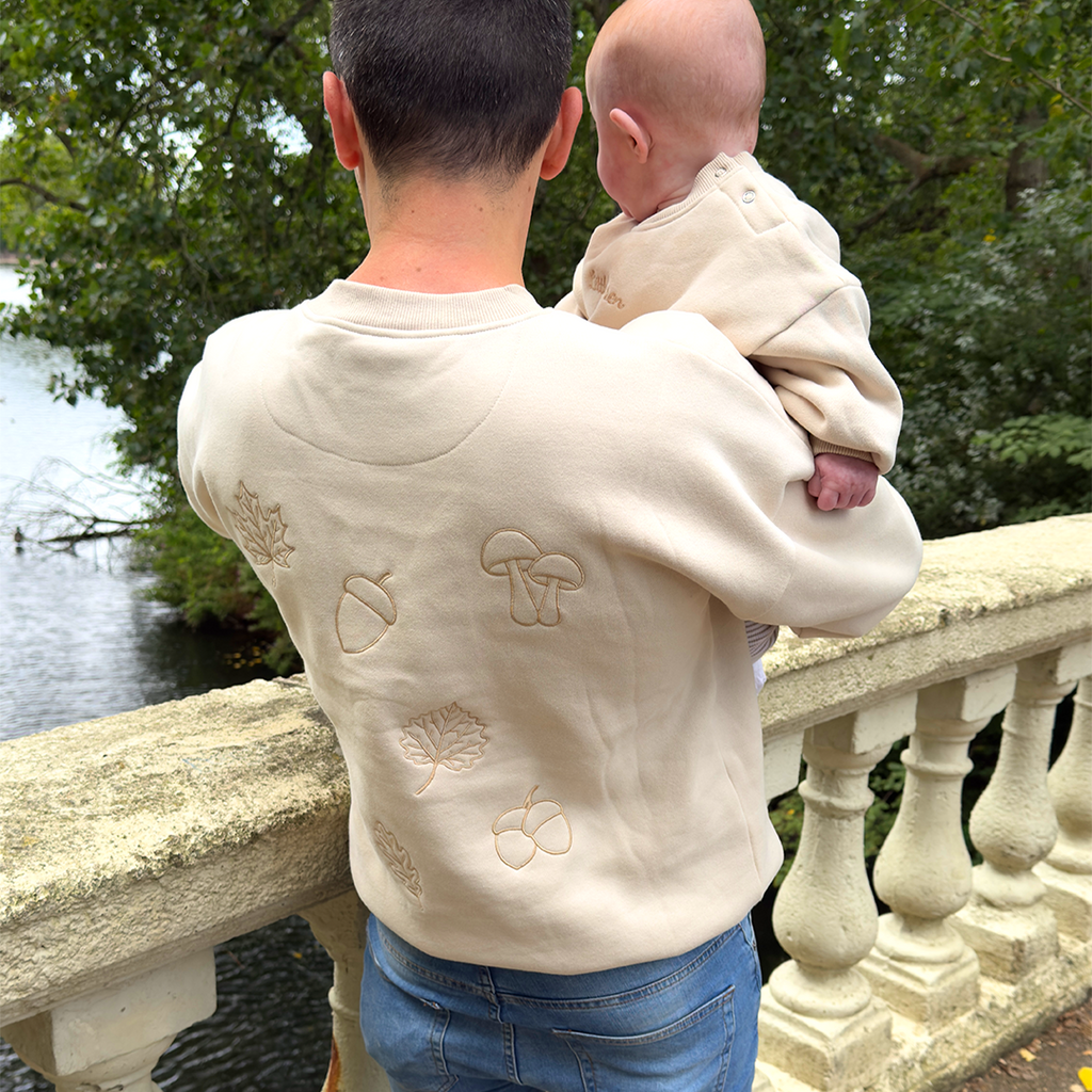 Person holding a baby wearing a cream-colored hoodie with embroidered designs by a body of water.