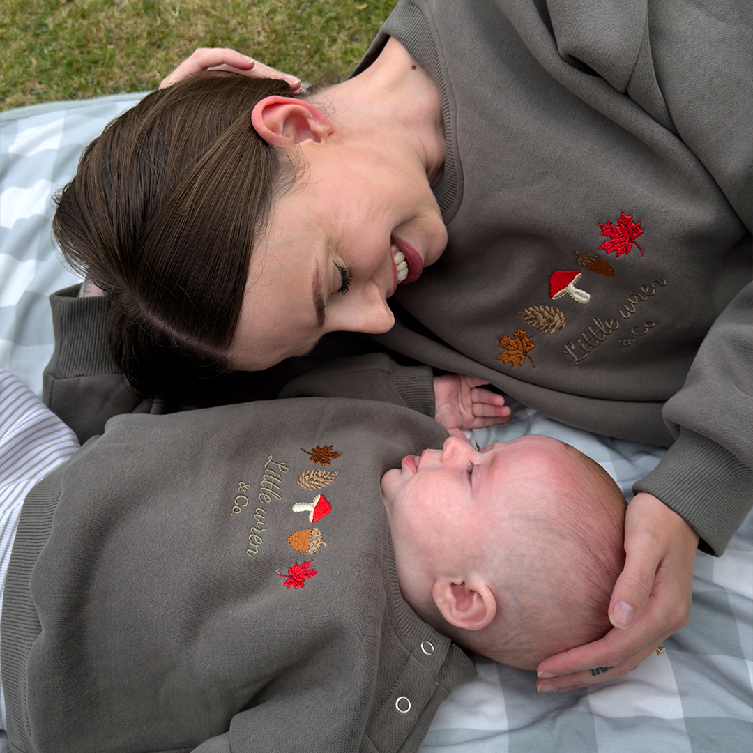 Woman and baby wearing matching sweatshirts with autumn-themed designs outdoors.