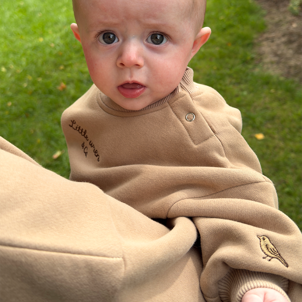 Baby in a beige outfit with text and a bird design, sitting on grass.
