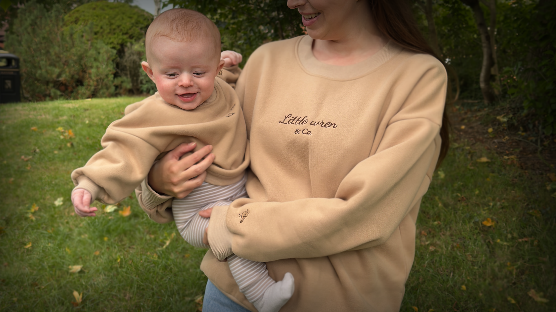 Woman holding a baby wearing matching beige sweatshirts with text in an outdoor setting.