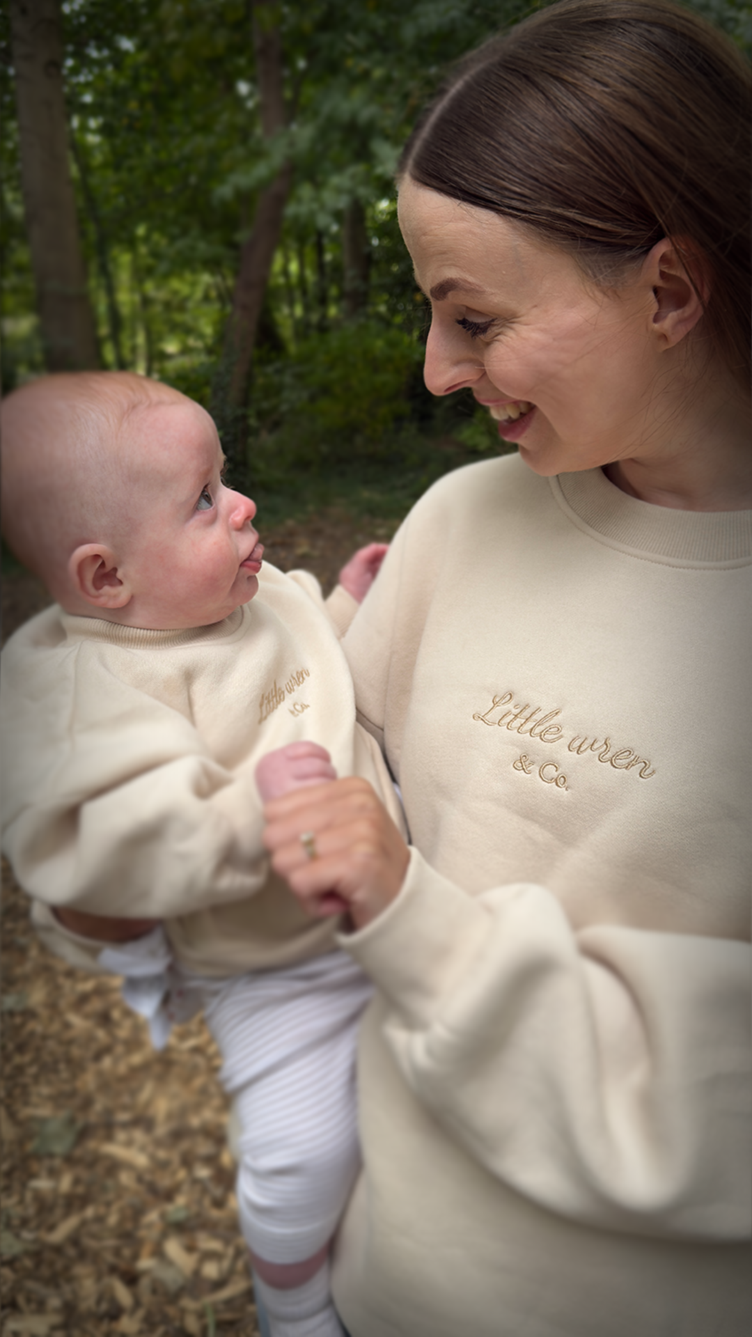Woman holding a baby outdoors, both wearing cream-colored outfits with 'Little Wren' branding.